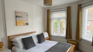 A neatly made bed with white linens and a grey throw, a wooden headboard, a bedside table with a lamp, a window with curtains, and a painting on the wall. - 59a Whitstable - 1 Bedroom Flat, Balcony & Parking (Whitstable)
