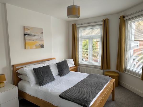 A neatly made bed with white linens and a grey throw, a wooden headboard, a bedside table with a lamp, a window with curtains, and a painting on the wall. - 59a Whitstable - 1 Bedroom Flat With Balcony (Whitstable)