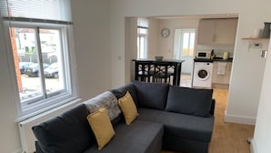 A modern living room with a grey sectional sofa, a dining table, and a kitchen in the background. - 59a Whitstable - 1 Bedroom Flat, Balcony & Parking (Whitstable)