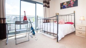 Un dormitorio con cama, escritorio, silla, ventana con cortinas, mesita de noche y un cuadro en la pared.
