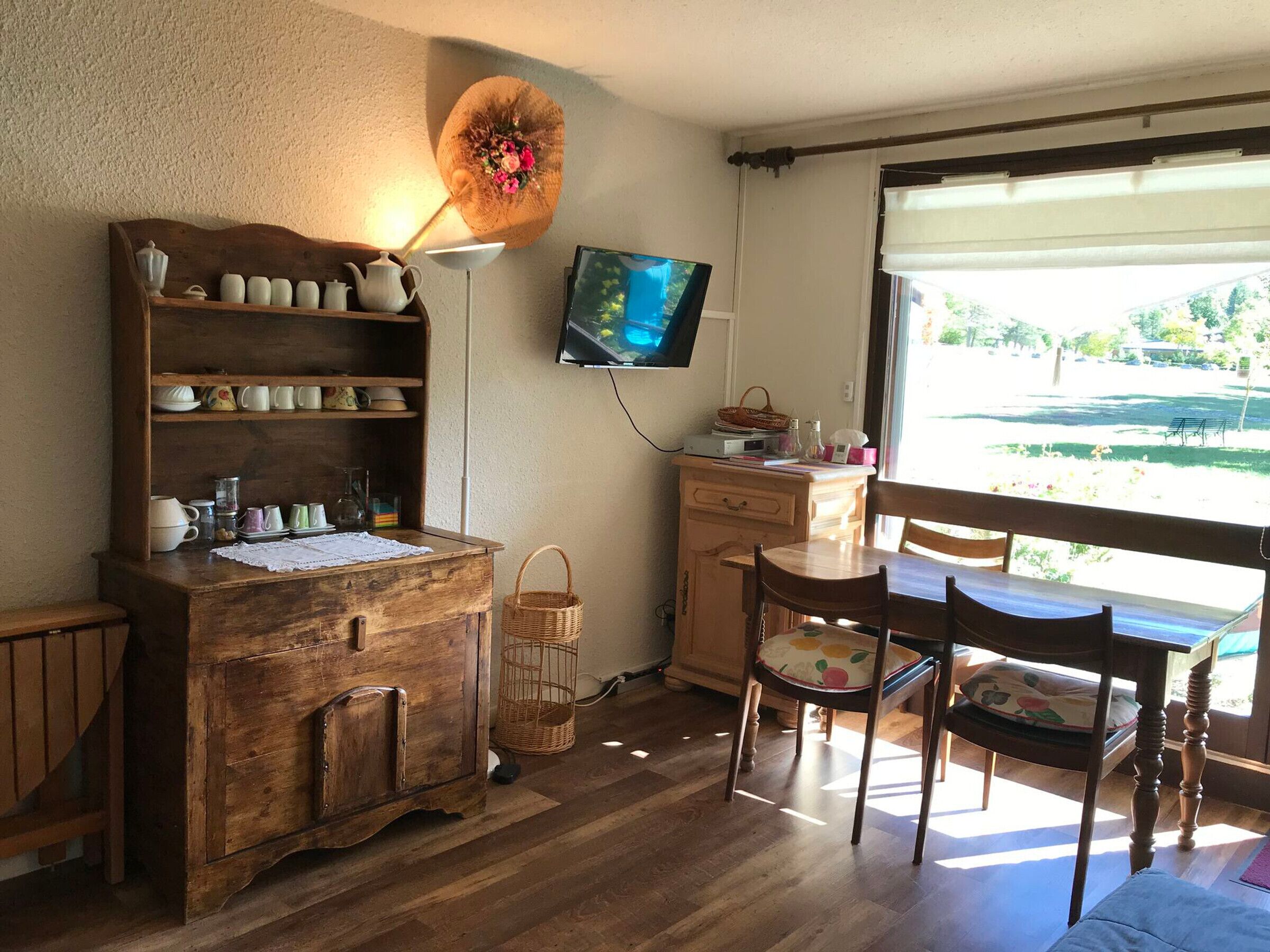 A cozy room with a wooden cabinet, a television, a desk, and a window with a view of greenery.