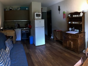 A compact kitchen with a microwave, oven, and dishwasher. There's a wooden dining table with chairs, a blue sofa, and a small cabinet with kitchenware. - Studio at the foot of the slopes in Corrençon (Corrençon-en-Vercors)