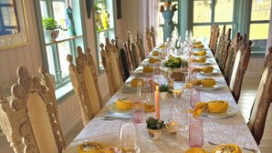A long dining table set with yellow bowls, pink glasses, and candles, surrounded by ornate chairs in a room with large windows and decorative plants.