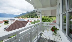 A balcony with a white railing, a table with a vase of flowers, and a view of houses and mountains in the background.