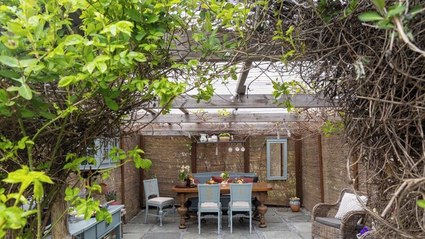 A covered outdoor seating area with a table and chairs, surrounded by plants and a wooden pergola.