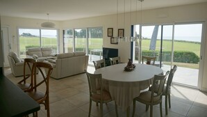 A spacious living area with a dining table, chairs, and a sofa. There is a large sliding glass door leading to a balcony with a view of the outdoors. - Ty Bihan, the "Little house" exceptional sea view! (Doëlan Clohars-Carnoët)