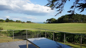 A modern outdoor terrace with a metal railing, a concrete table, and a view of a grassy field and trees. - Ty Bihan, the "Little house" exceptional sea view! (Doëlan Clohars-Carnoët)