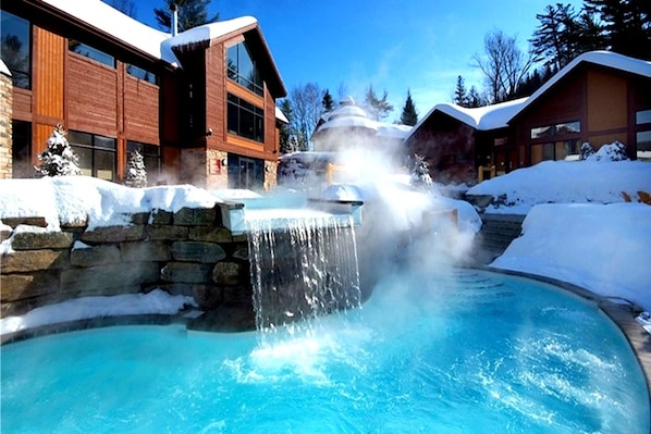 A pool with a waterfall feature surrounded by snow-covered buildings.