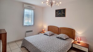 Een slaapkamer met een bed, nachtkastje, lamp, raam met zonwering, radiator, plafondlamp en een lijst met een afbeelding aan de muur.
