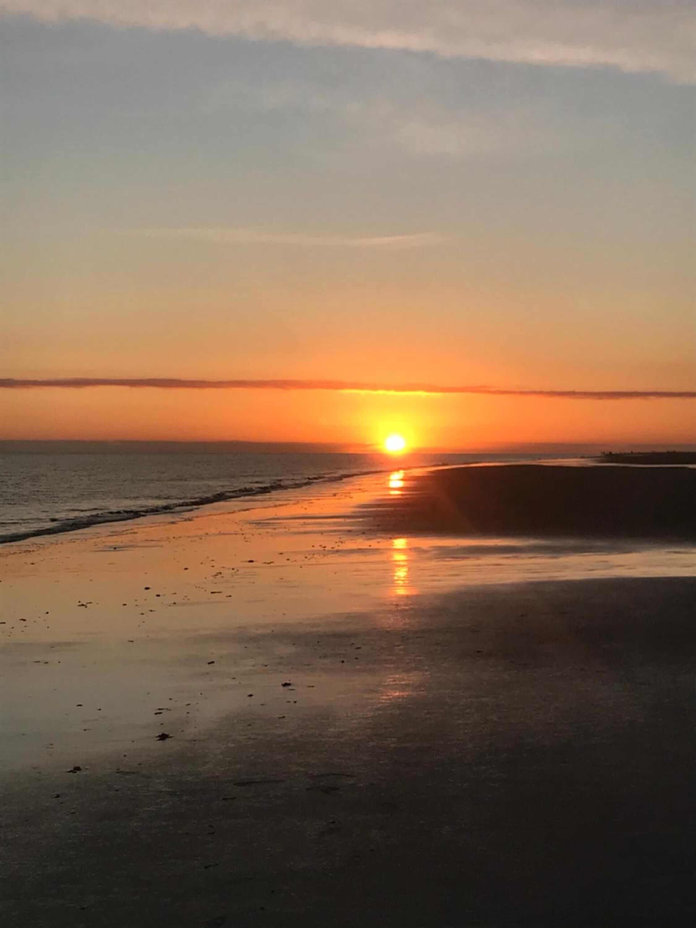 A beach at sunset with the sun low on the horizon, reflecting on the wet sand.