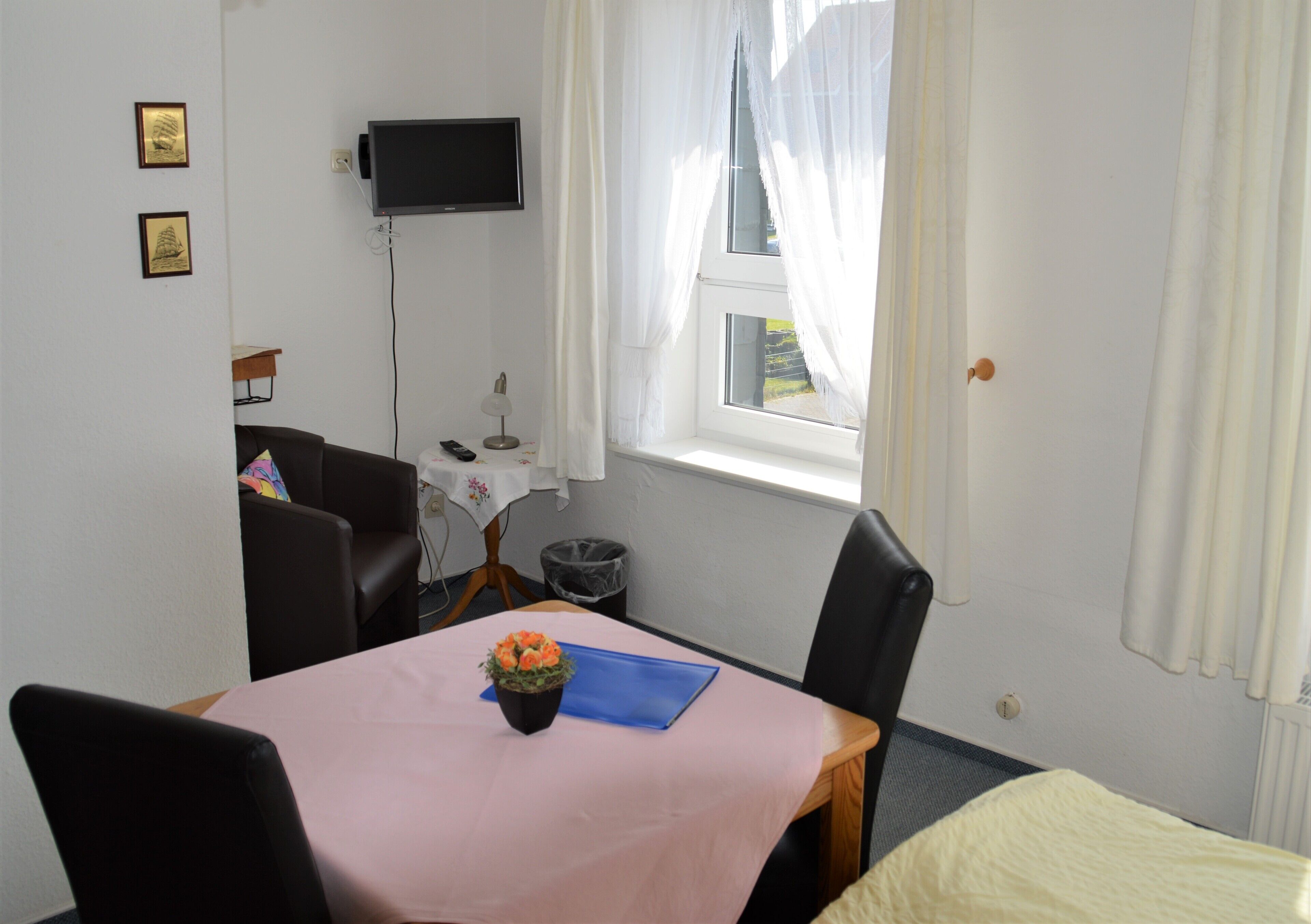 A hotel room with a bed, a desk with a computer, a chair, a small table with a flower, and a window with curtains.