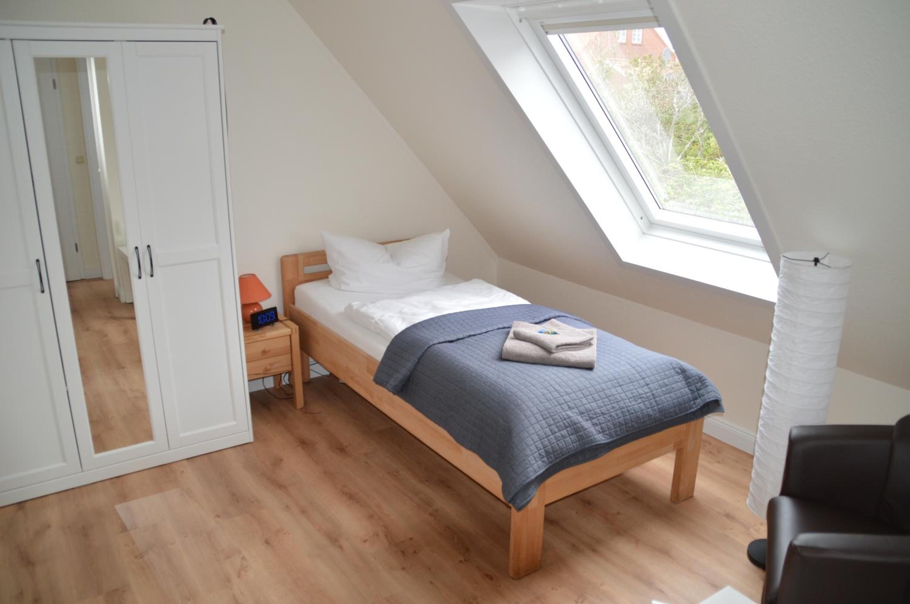 A bedroom with a sloped ceiling, a bed with a blue quilt, a nightstand with a lamp, and a window with a view of greenery.
