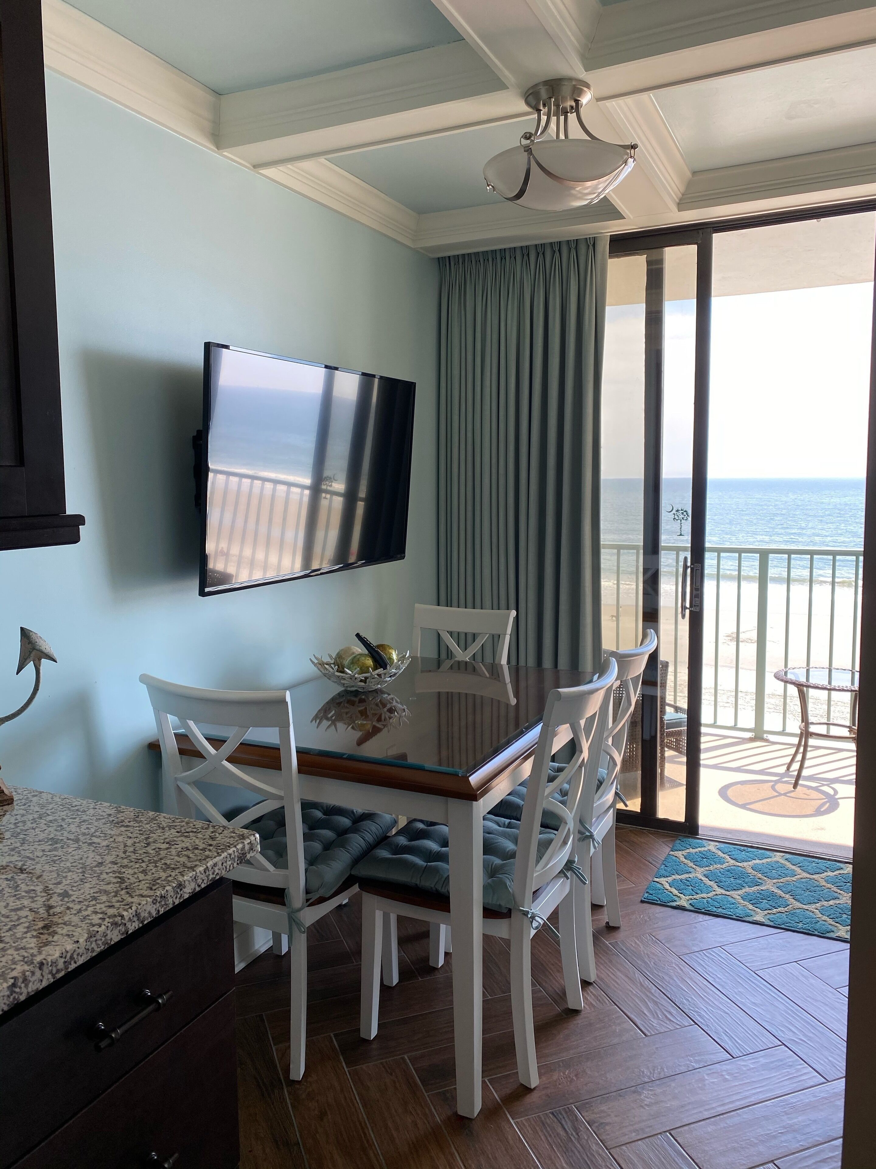 A dining area with a TV mounted on the wall, a table with chairs, and a view of the ocean through a sliding glass door.