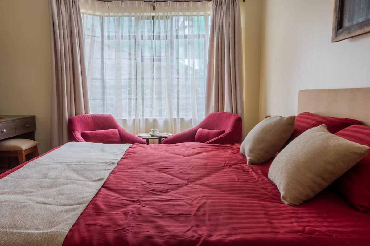 A hotel room with a large bed, two red armchairs, a small table with a cup, and a window with sheer curtains.