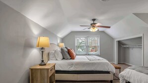 Un dormitorio con techo inclinado, una cama con ropa de cama blanca, una mesita de noche de madera, una lámpara de pie y un ventilador de techo.