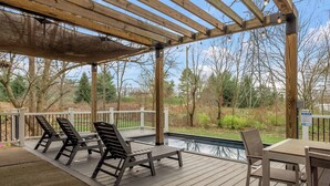 Una terraza de madera con mobiliario exterior, una piscina y vistas a árboles y un edificio al fondo.