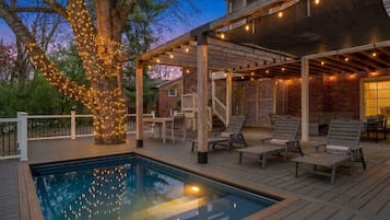 A poolside area with a wooden deck, lounge chairs, and a pergola with string lights.