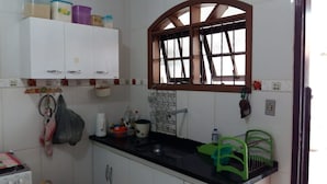 A kitchen with white cabinets, a black countertop, a window with a wooden frame, and a hanging rack with dishes. - Casa de Praia Cordeirinho (Marica)