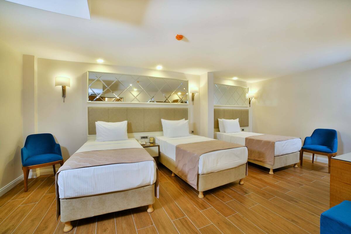 A hotel room with three beds, wooden flooring, and a large mirror with geometric patterns.