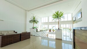 Un hall d’entrée spacieux avec un plafond haut, de grandes fenêtres, un coin salon avec un canapé et des fauteuils, et deux plantes en pot.