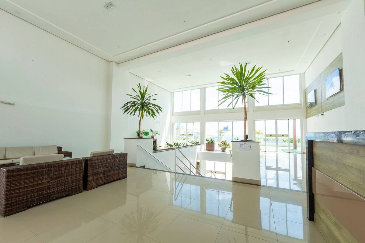 A spacious lobby with a high ceiling, large windows, a seating area with a sofa and chairs, and two potted plants.