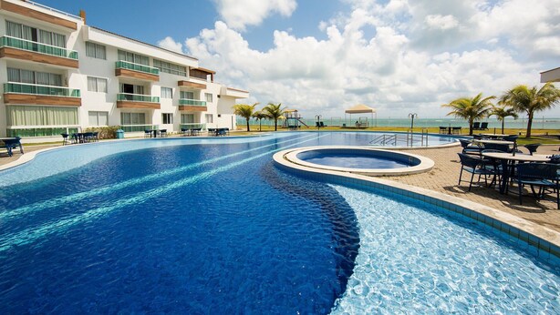 Une piscine au design incurvé, entourée de chaises longues et de tables, et un bâtiment avec des balcons en arrière-plan.