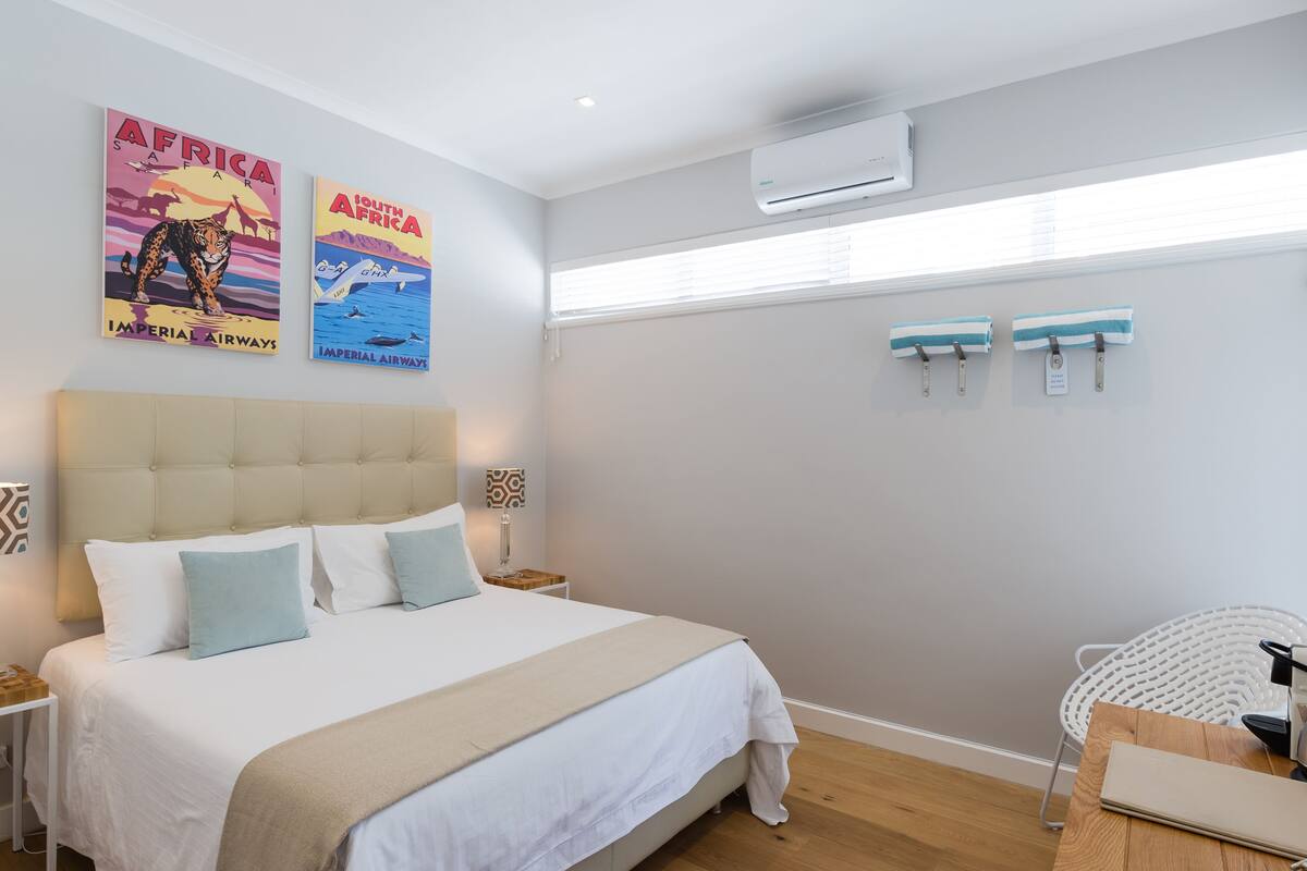 A bedroom with a large bed, two wall-mounted air conditioning units, a window with blinds, and two towels hanging on hooks.