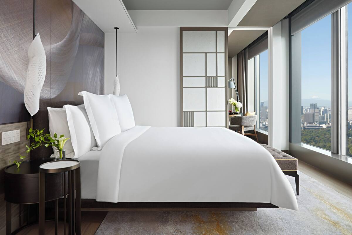 A modern bedroom with a large bed, a bedside table with a plant, a chair, and a view of the city through large windows.