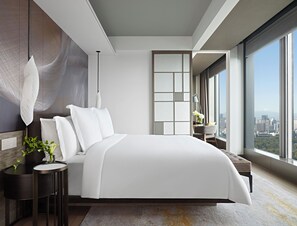 A modern bedroom with a large bed, a bedside table with a plant, a chair, and a view of the city through large windows. - Four Seasons Hotel Tokyo at Otemachi (Tokyo)