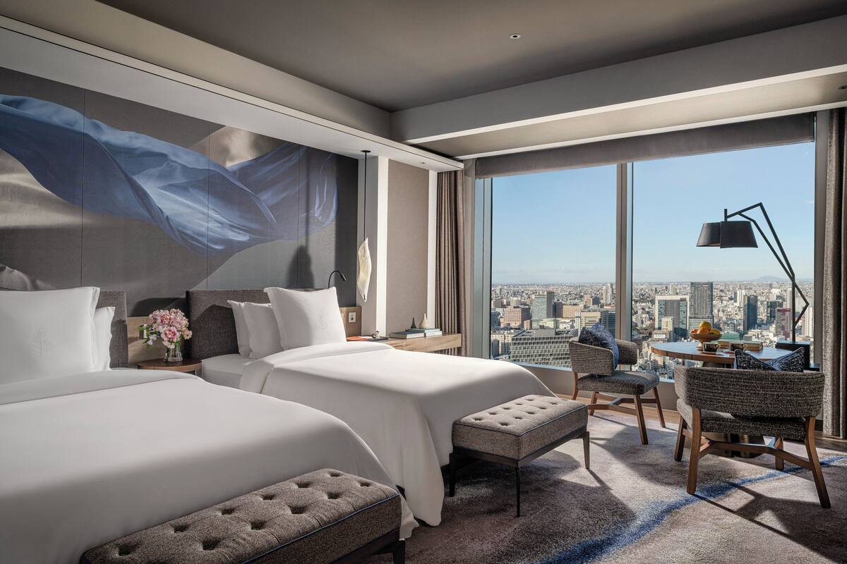A modern hotel room with two beds, a large window with a city view, a dining area, and a view of the cityscape.