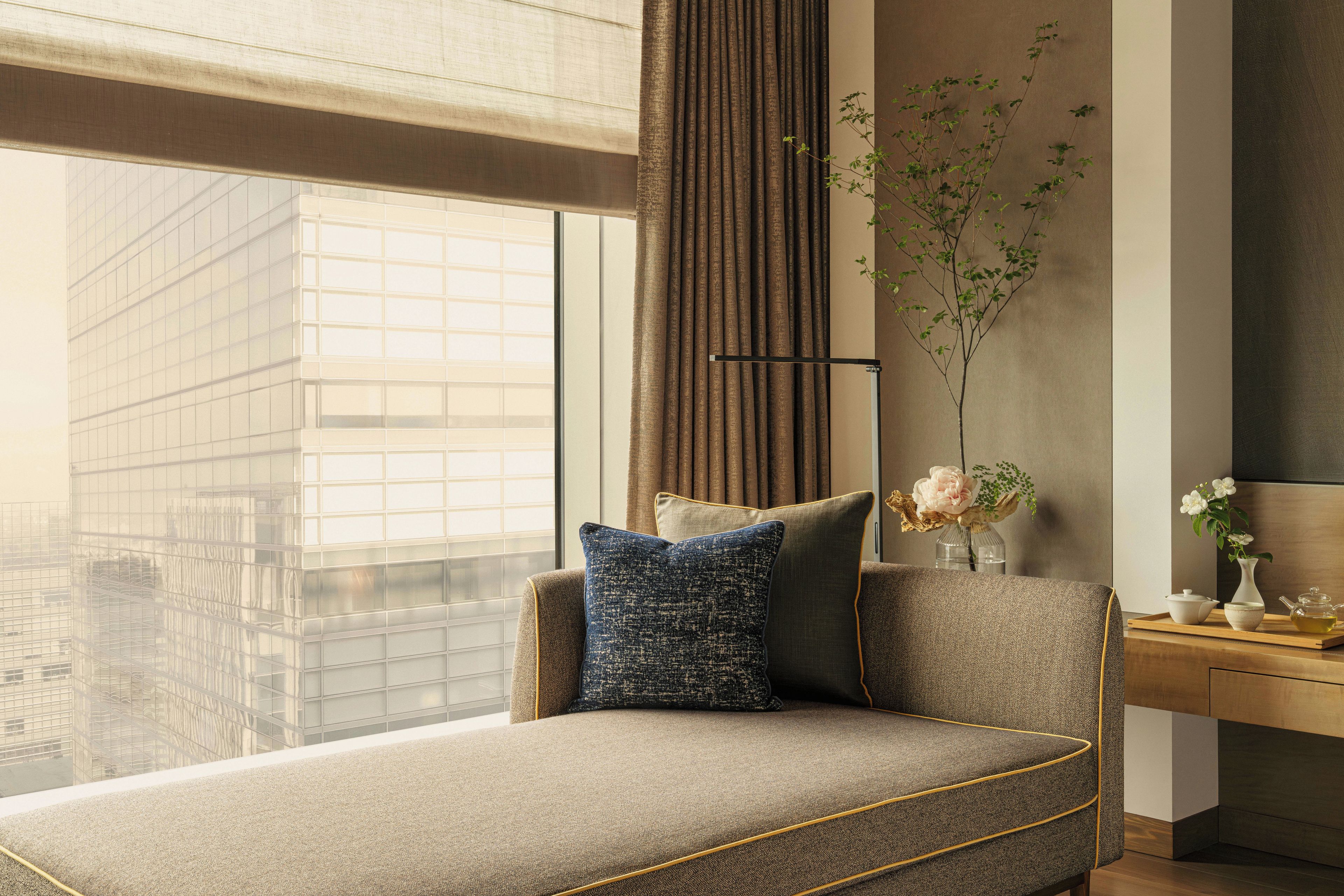Un salón moderno con un sofá, un florero con flores y vistas a un edificio a través de grandes ventanales.
