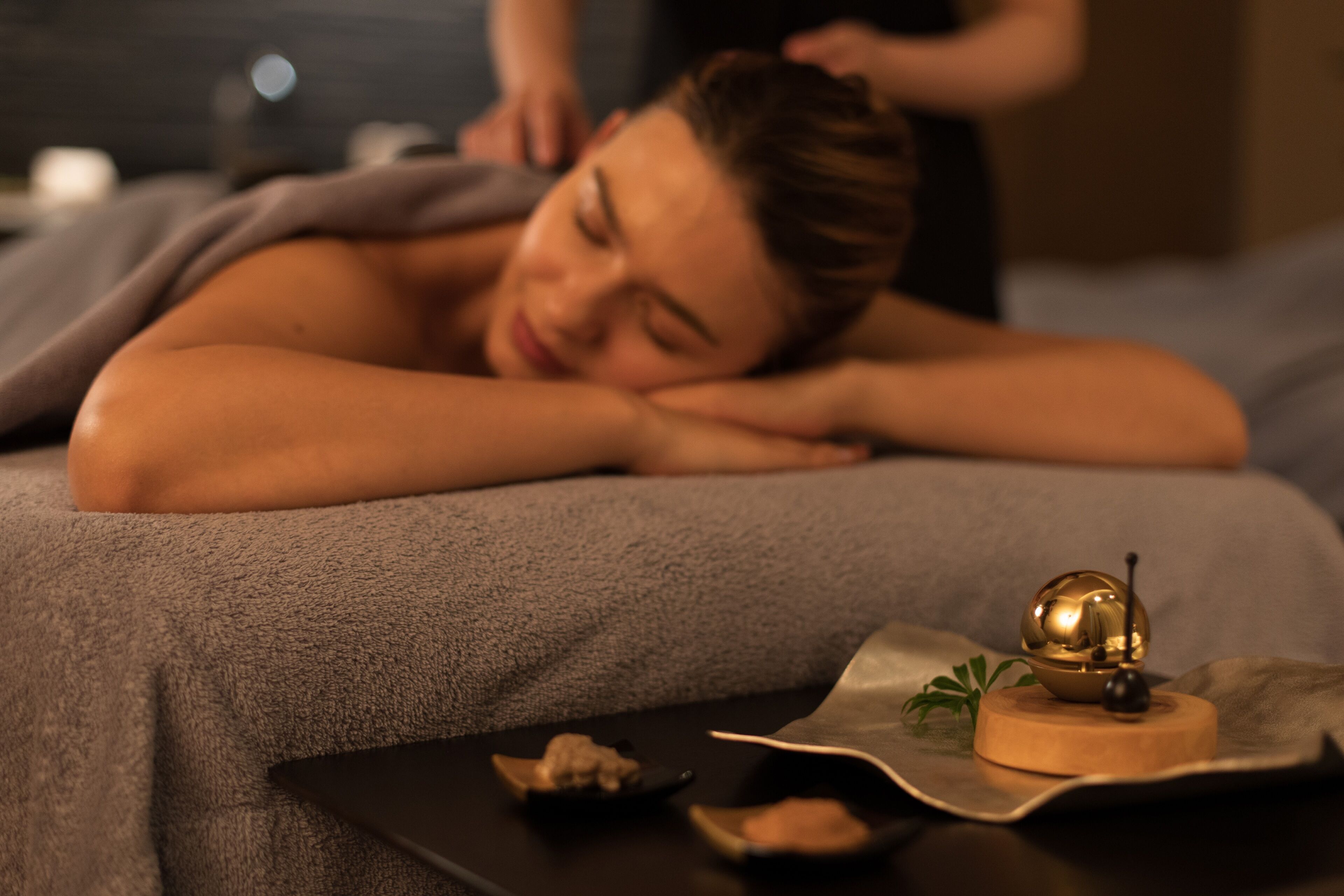 A person lying on a massage table with a massage therapist attending to them. There is a small wooden table with a decorative item and a book on it.