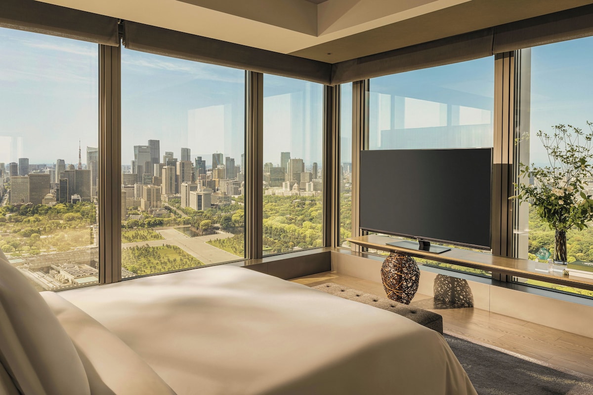 A modern hotel room with a large bed, a flat-screen TV, and a city view through floor-to-ceiling windows.
