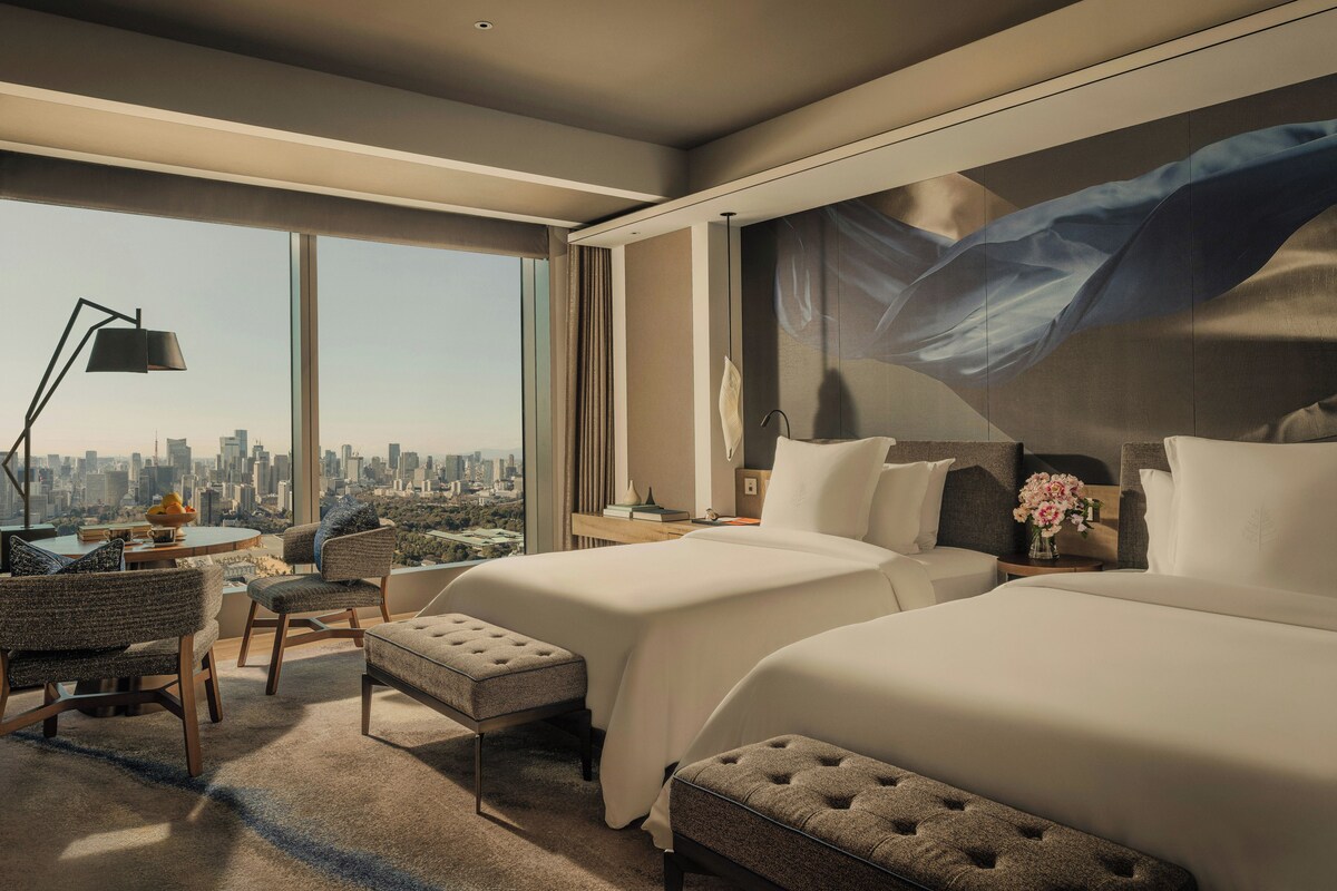 A modern hotel room with a city view, two beds, a desk, and a large abstract painting on the wall.