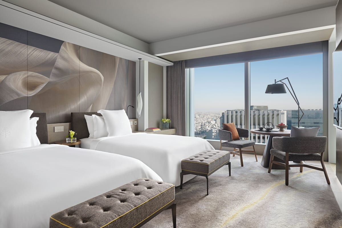 A modern hotel room with two large beds, a sitting area with a chair and table, and a large window offering a city view.