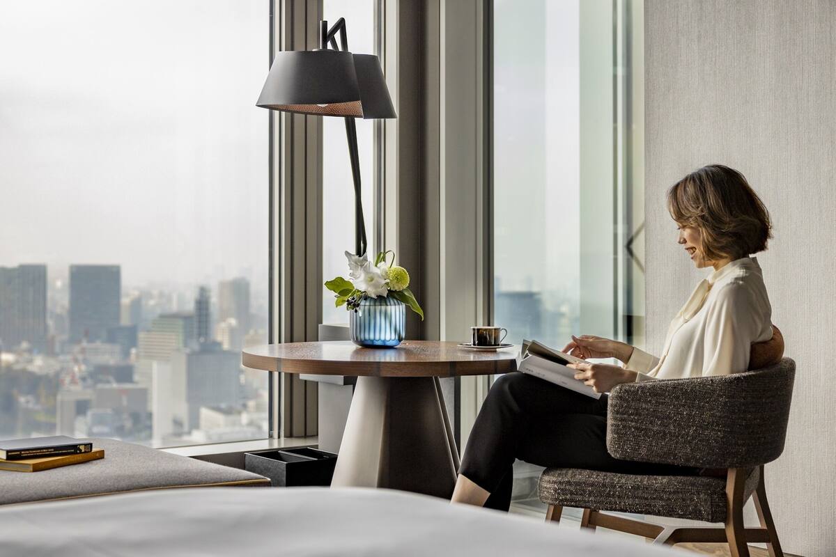 A modern hotel room with a large window offering a city view, a round table with a vase of flowers, and a person reading a book.