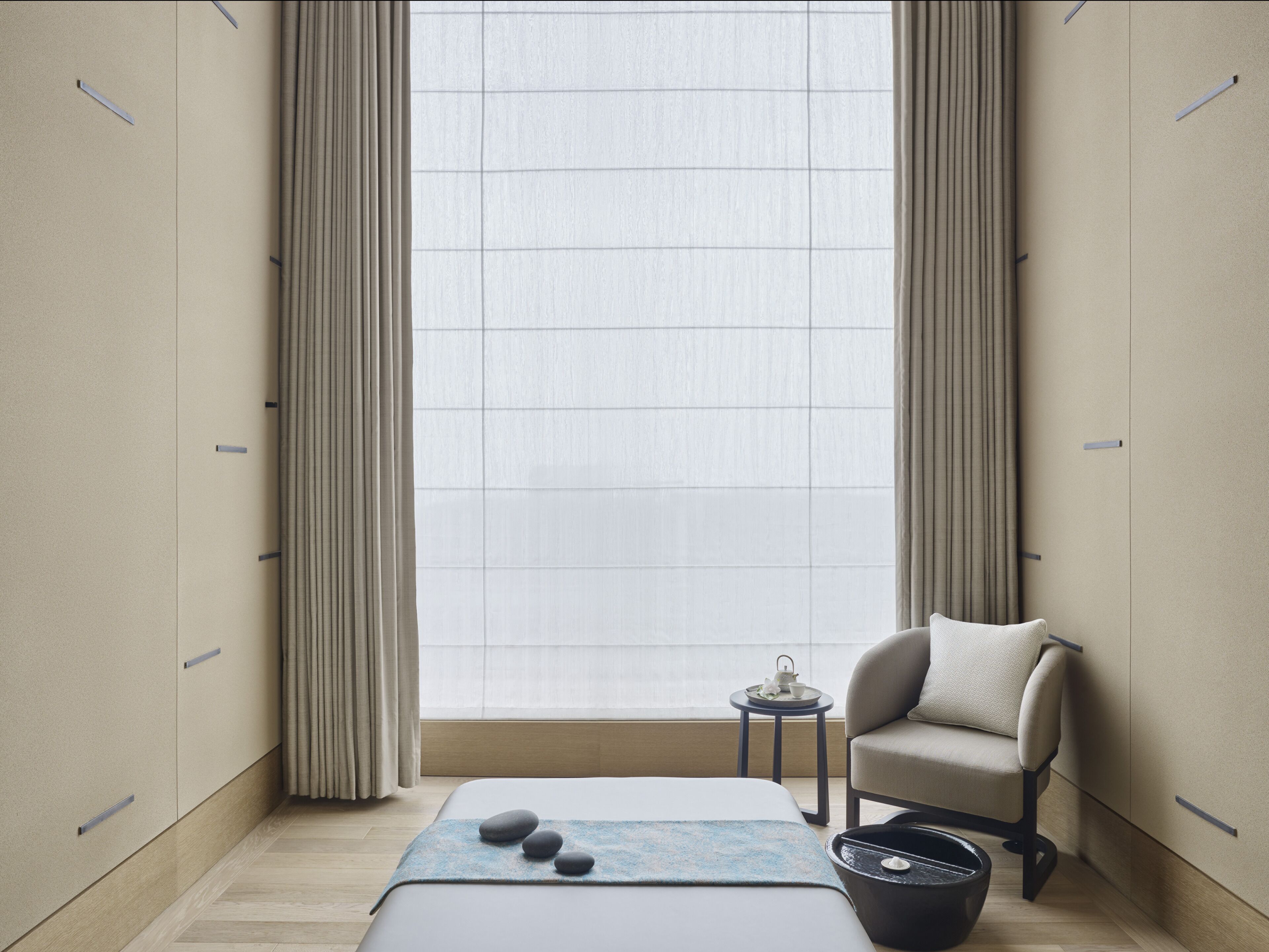 A modern bedroom with a large window, a bed with a blue blanket, a chair, and a small table with a teapot.