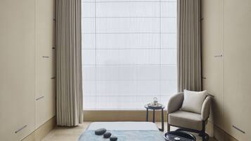 A modern bedroom with a large window, a bed with a blue blanket, a chair, and a small table with a teapot.