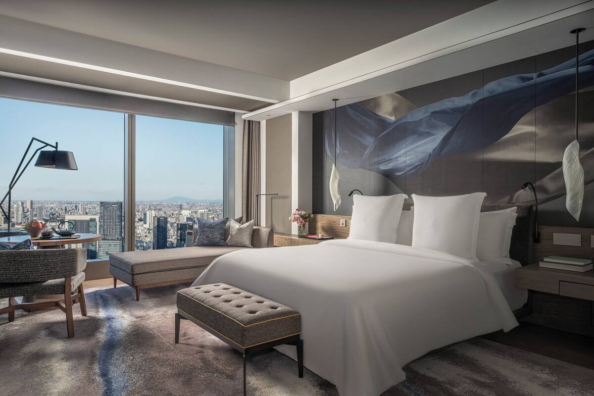 A modern hotel room with a large bed, a desk with a lamp, a view of the city, and a large window.