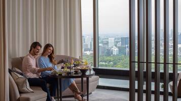 Una pareja sentada en un sofá en una sala de estar moderna con vista a la ciudad, una mesita con bebidas y un ventanal con persianas.