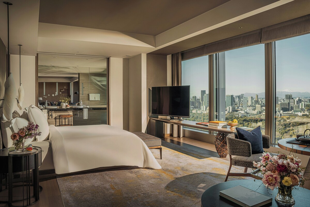 A modern hotel room with a large bed, a desk with a TV, and a view of a cityscape.