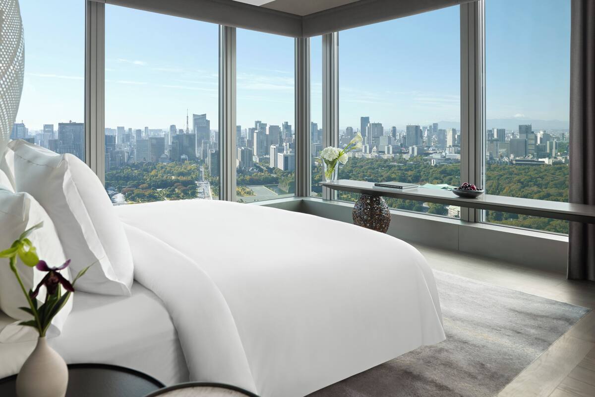 A modern bedroom with a large bed, a vase with flowers, and a view of a cityscape through large windows.