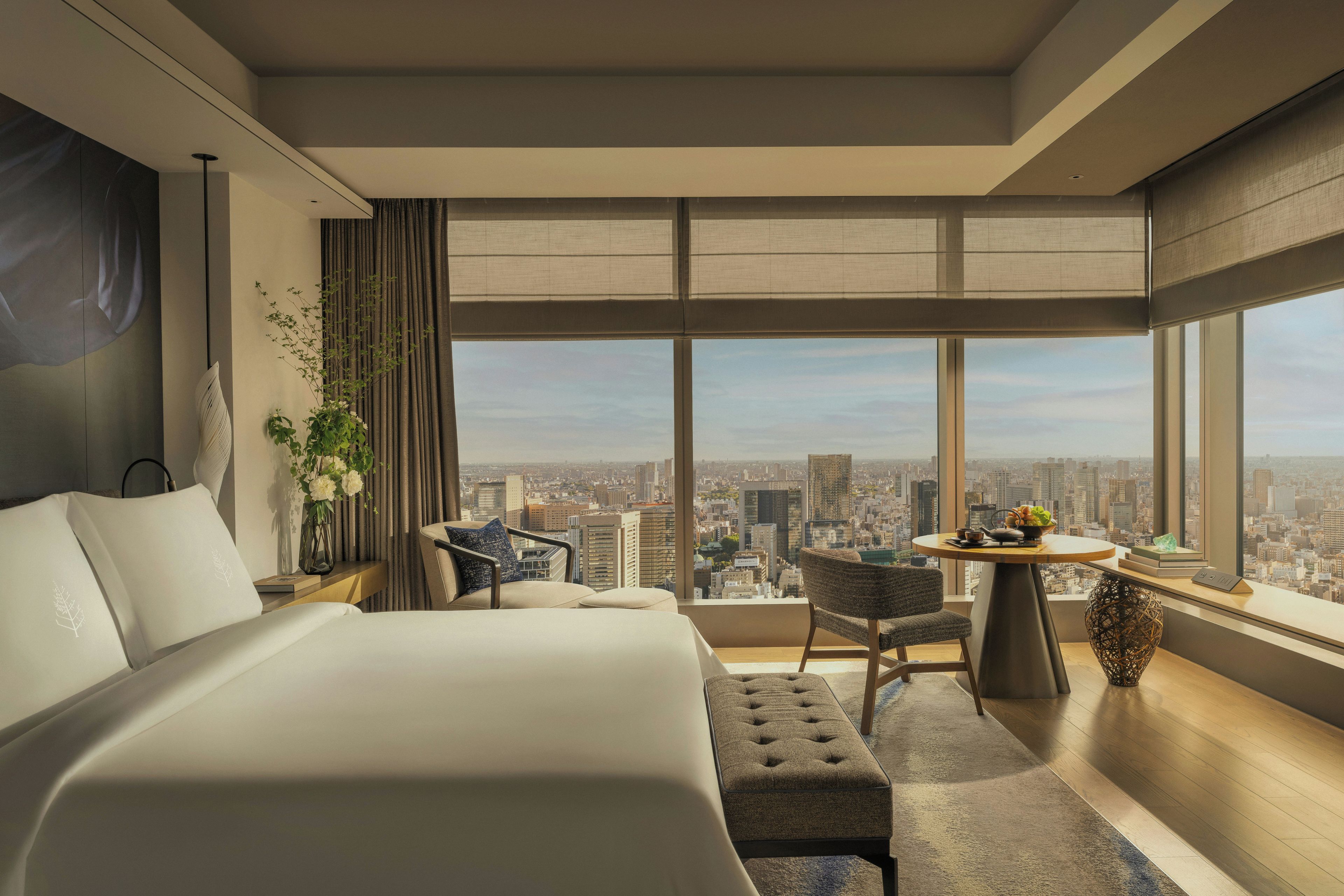 A modern hotel room with a large bed, a seating area with a table, and a city view through large windows.