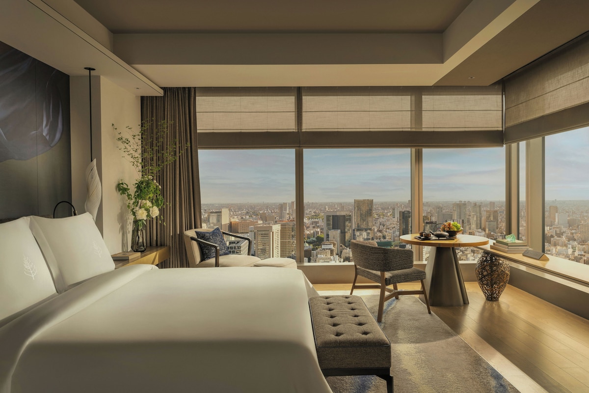 A modern hotel room with a large bed, a seating area with a table, and a city view through large windows.