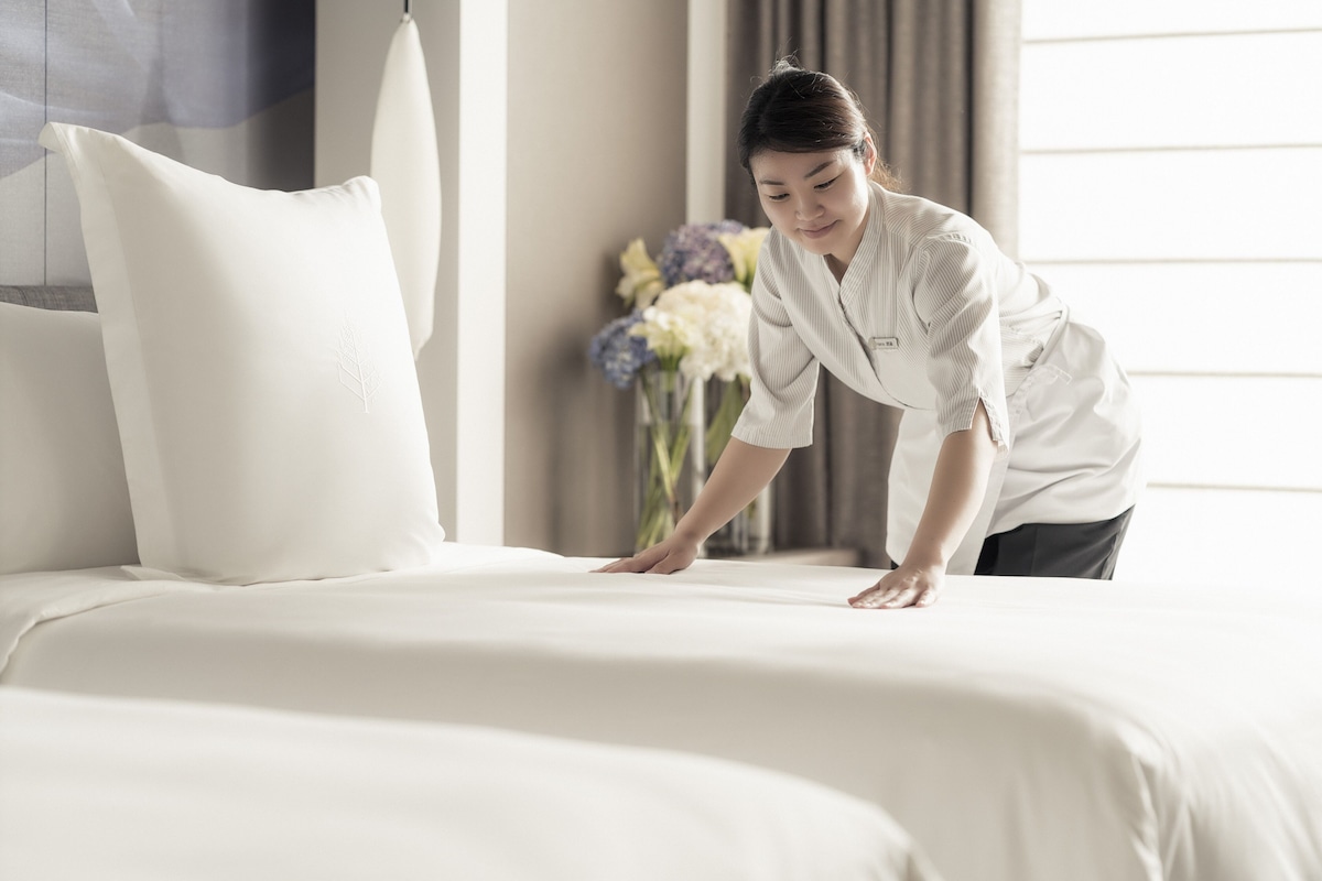 A hotel room with a neatly made bed, a large pillow, and a vase of flowers in the background.