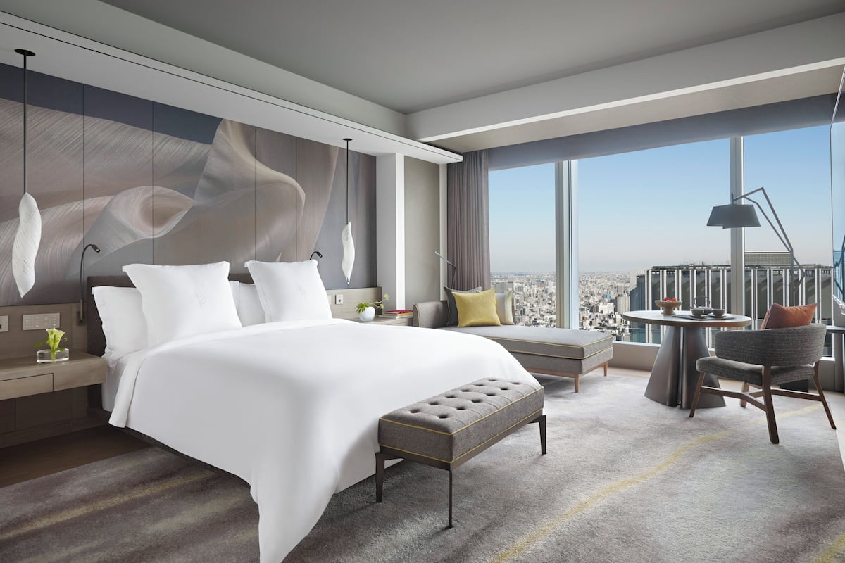 A modern hotel room with a large bed, a seating area with a view, and a dining table with a cityscape view.