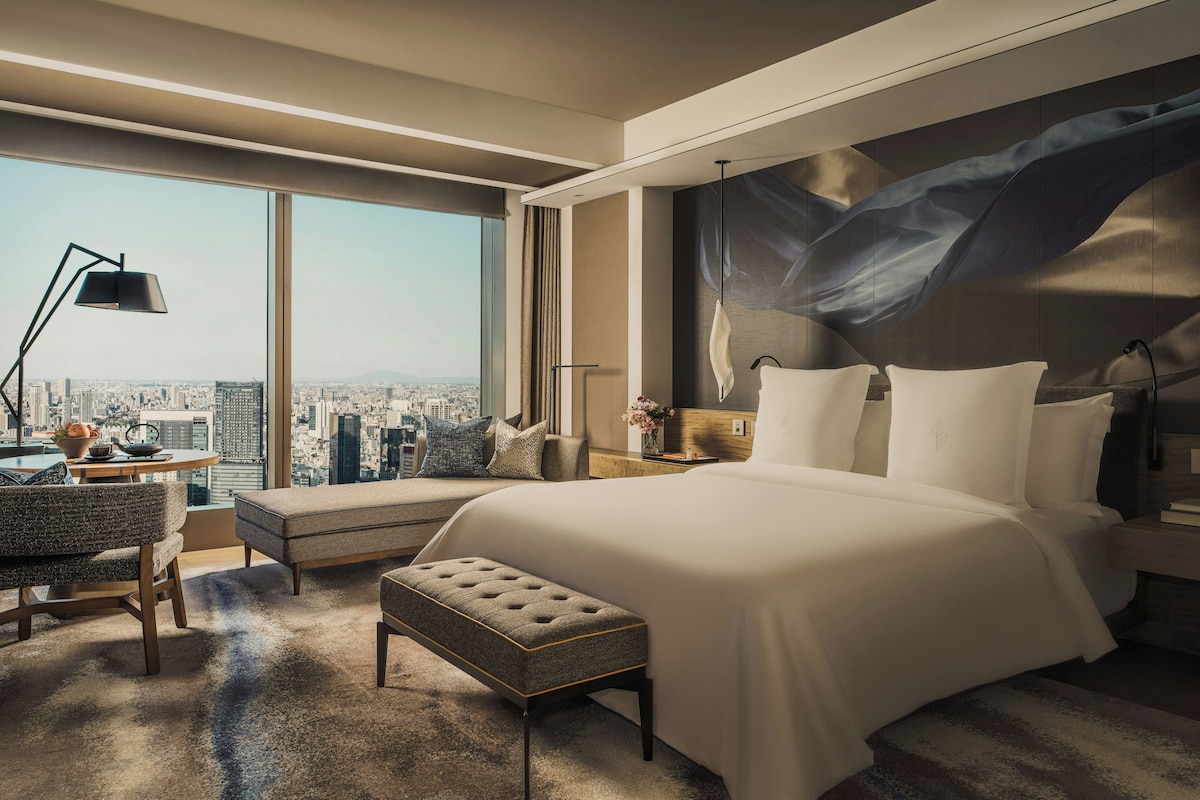A modern hotel room with a large bed, a desk, a view of the city, and a cityscape through the window.