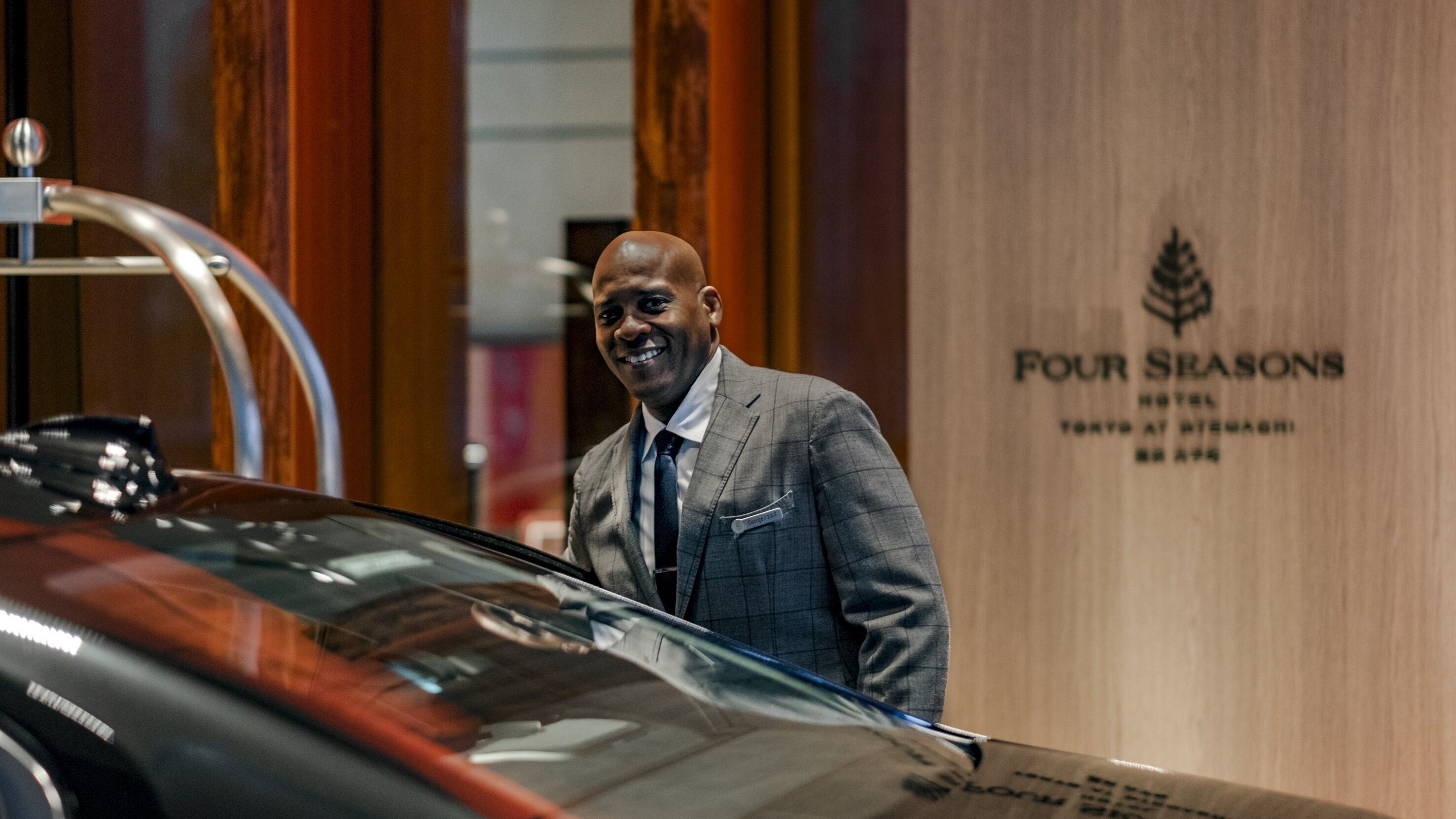 A man in a suit standing next to a car in front of a wooden wall with the text "Four Seasons Hotel".