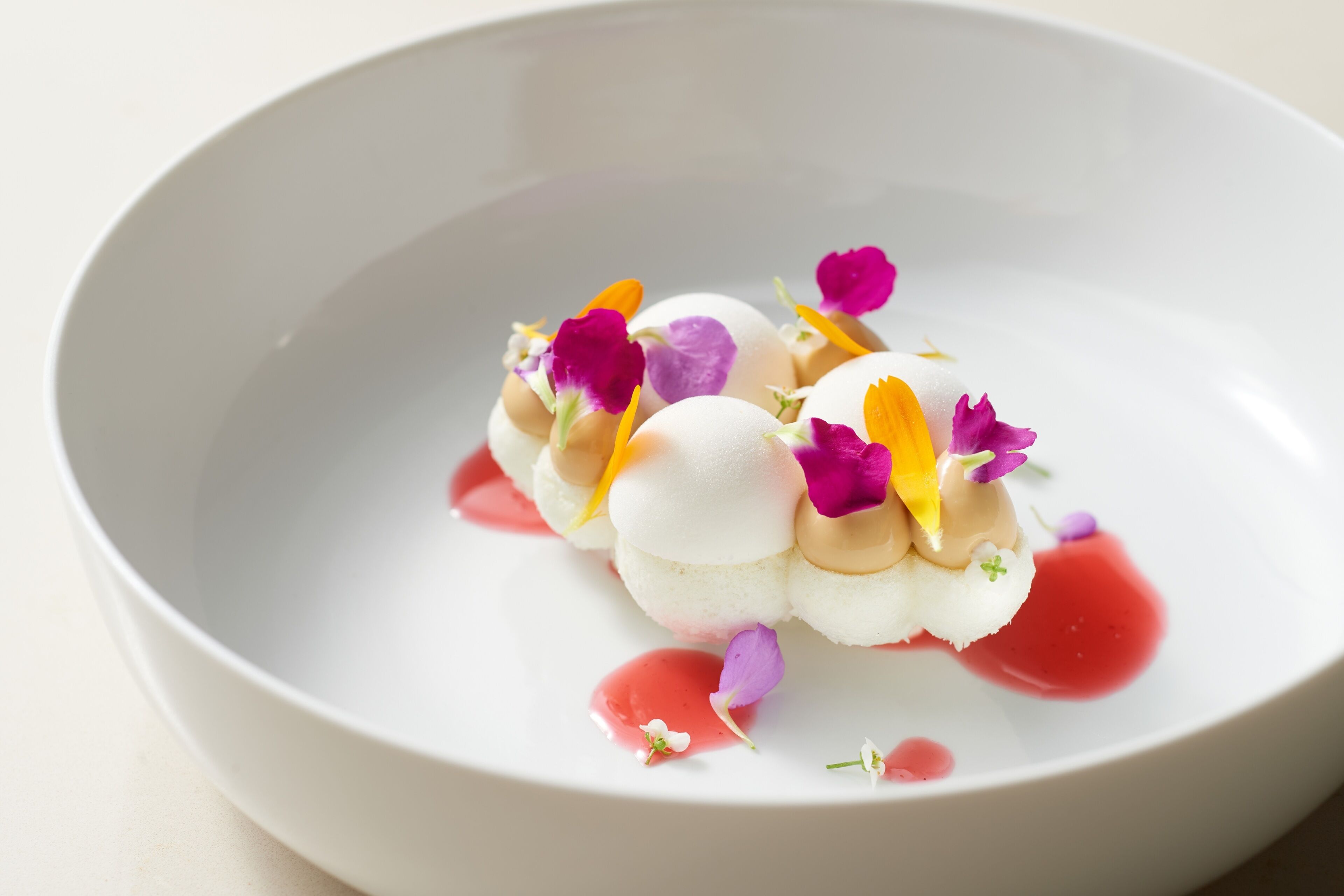 A white plate with a dessert consisting of spherical white and beige components, garnished with purple and yellow flowers and a red sauce.