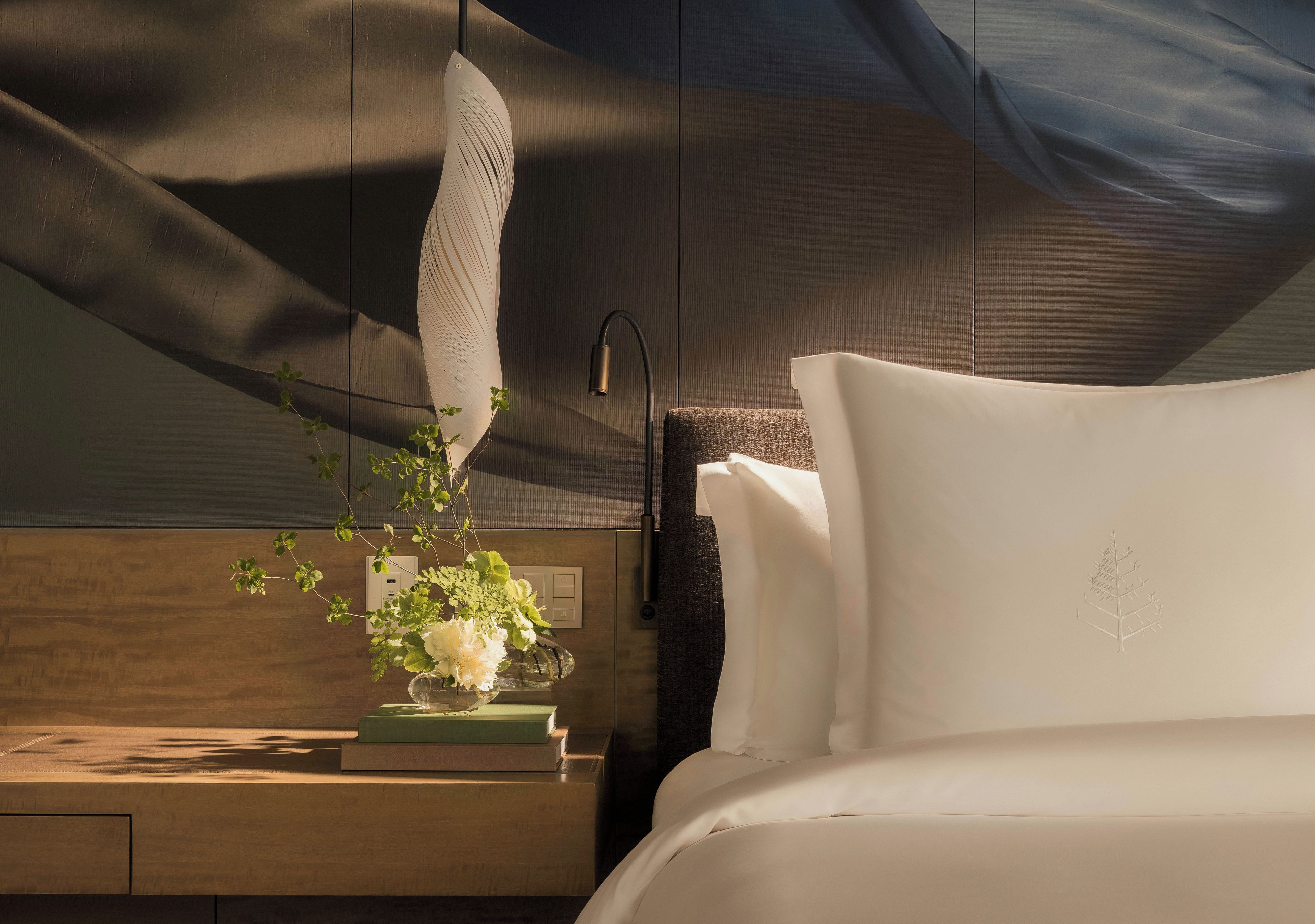 A modern hotel room with a bed, a bedside table with a lamp, and a wall-mounted control panel.